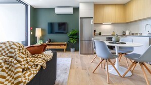 Una habitación de hotel moderna con sofá, una mesa de comedor con sillas, una zona de cocina con refrigerador y un televisor de pantalla plana montado en la pared.