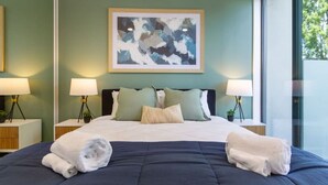 Una cama bien hecha con toallas blancas, un edredón azul oscuro y almohadas verdes y blancas. Hay dos mesitas de noche con lámparas y pequeñas macetas. Encima de la cama cuelga un cuadro abstracto enmarcado.