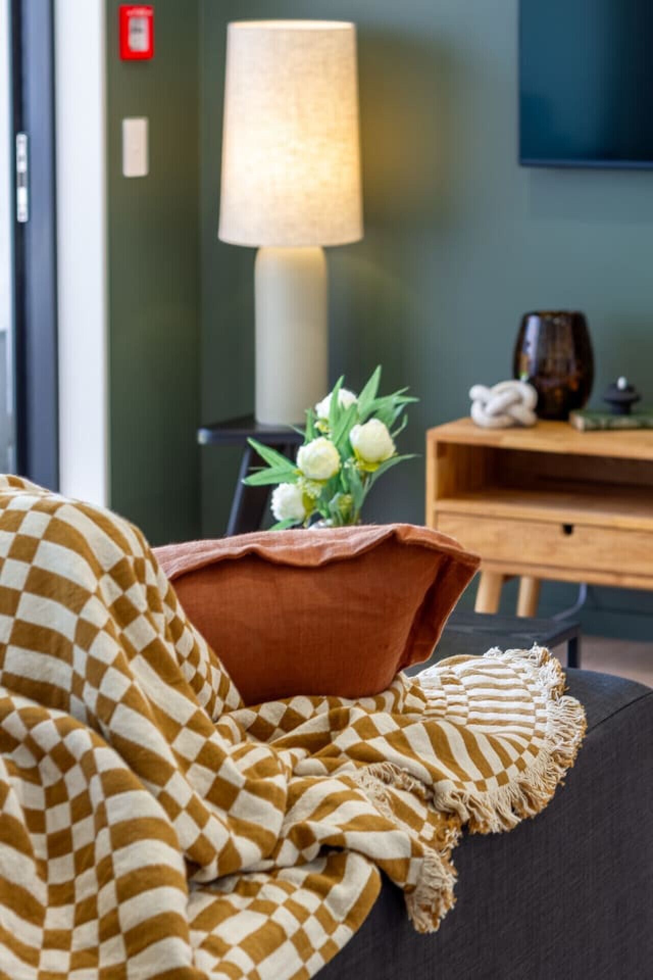 A cozy room with a checkered blanket, a brown pillow, and a vase with white flowers on a dark sofa. A lamp and a wooden side table with a vase and a candle are in the background.