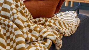 Una habitación acogedora con una manta a cuadros, un cojín marrón y un jarrón con flores blancas sobre un sofá oscuro. Una lámpara y una mesita de noche de madera con un jarrón y una vela se ven al fondo.