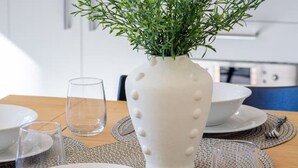 Una tavola imbandita con piatti, ciotole e bicchieri bianchi, un vaso bianco con piante verdi e una cucina sullo sfondo.