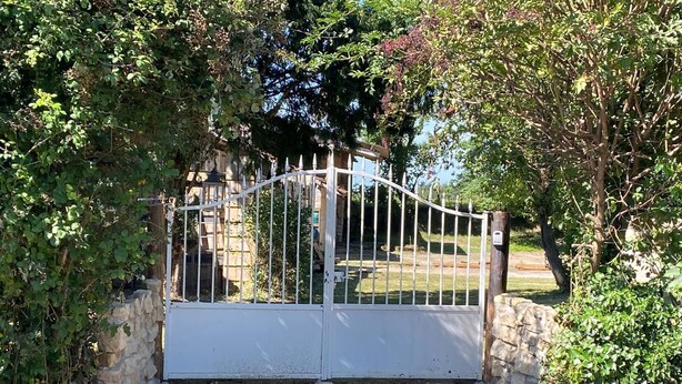 Una entrada tancada amb una reixa de metall blanca, murs de pedra i vegetació.