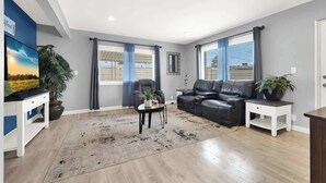 Ett modernt vardagsrum med en svart lädersoffa, ett vitt soffbord och en tv som står på ett tv-bord. Det finns två fönster med blå gardiner, en krukväxt och ett litet sidobord.