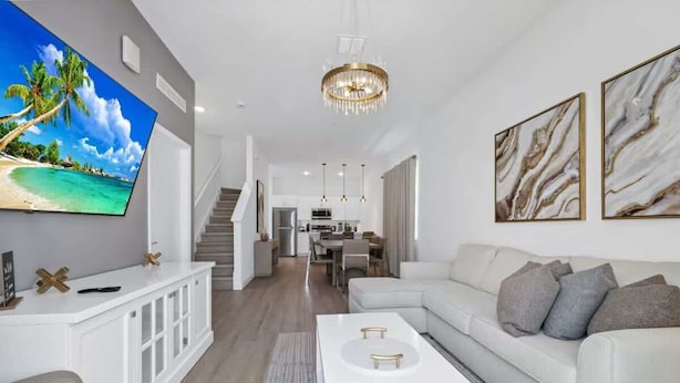 Ein modernes Wohnzimmer mit einem weißen Sofa, einem Couchtisch und einem an der Wand montierten Fernseher. Eine Treppe führt zu einer oberen Ebene, und ein Kronleuchter hängt von der Decke.