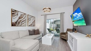 Ein modernes Wohnzimmer mit einem weißen Sofa, einem Couchtisch und einem an der Wand befestigten Fernseher. Es gibt einen Balkon mit einer Glastür und Blick auf den Ozean.
