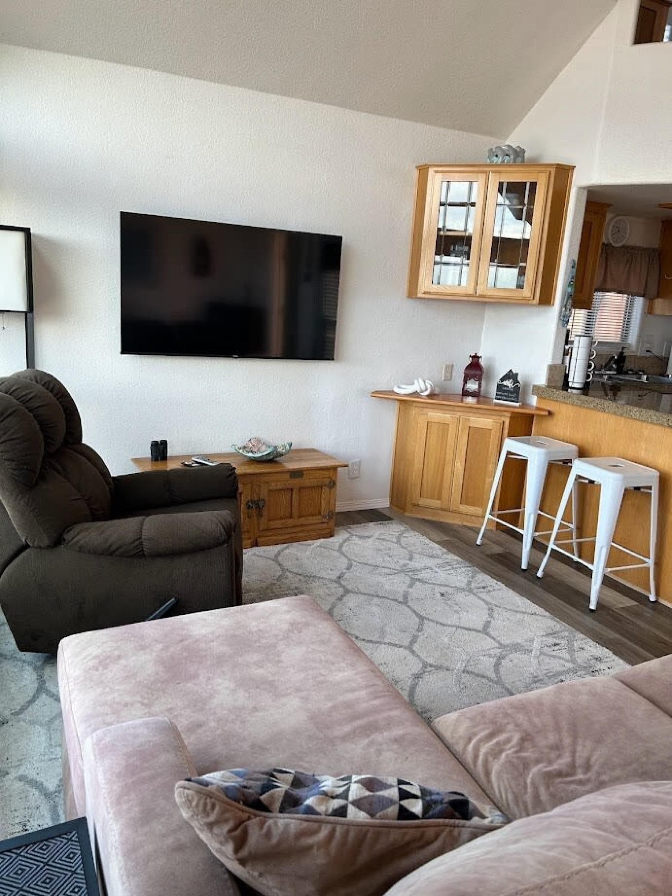 A living room with a sofa, a TV, a wooden cabinet, and a kitchen area with stools.