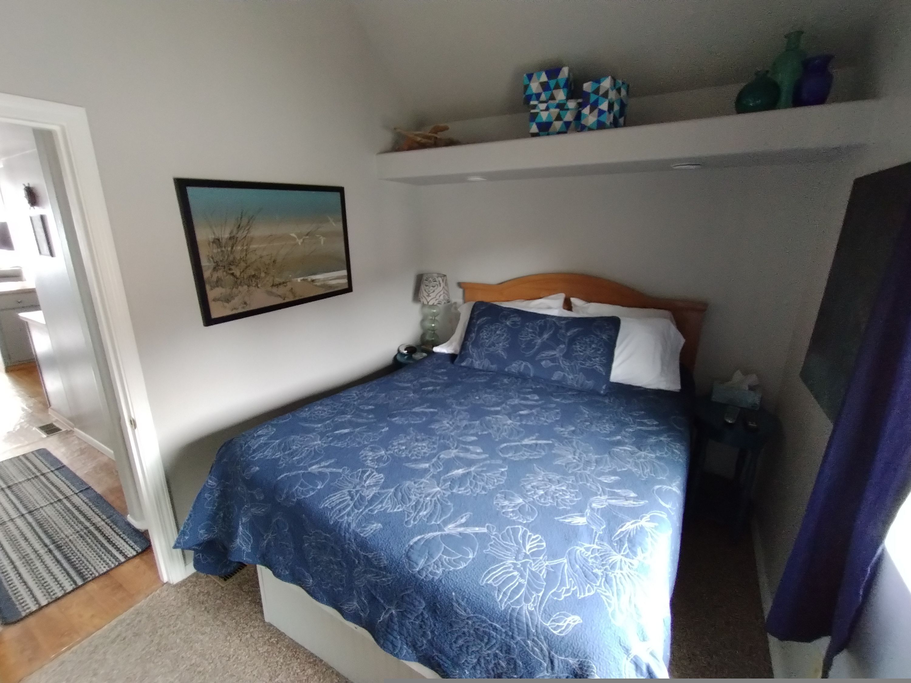 Une chambre à coucher comprenant un lit, une table de chevet, une lampe, un tableau encadré et une étagère avec des objets décoratifs.