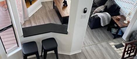Ett modernt kök med en svart bänkskiva och två barstolar, en matplats med ett träbord och stolar samt en mysig sittgrupp med en soffa och kuddar.