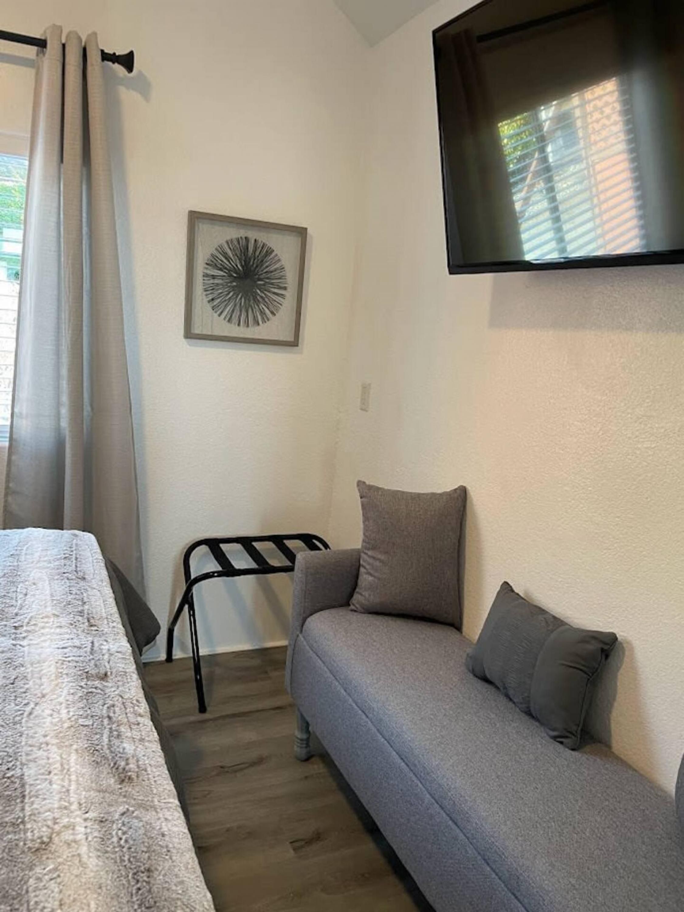 A room with a bed, a gray sofa with pillows, a black metal chair, a framed abstract art piece, and a mounted TV.