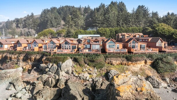 Una hilera de cabañas de madera con ventanas amplias, frente a un terreno rocoso y un bosque denso.
