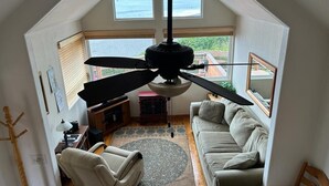 Ein geräumiges Wohnzimmer mit Deckenventilator, einer Couch, einem Sessel und Blick ins Freie durch große Fenster.
