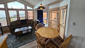 A cozy living room with a sofa, coffee table, and dining table with chairs. There are large windows and a ceiling fan.