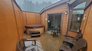A wooden deck with a hot tub, two chairs, and a glass door leading to a balcony.