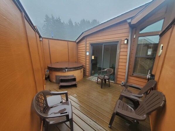 Una terraza de madera con un jacuzzi, dos sillas y un ventanal que da a un balcón.