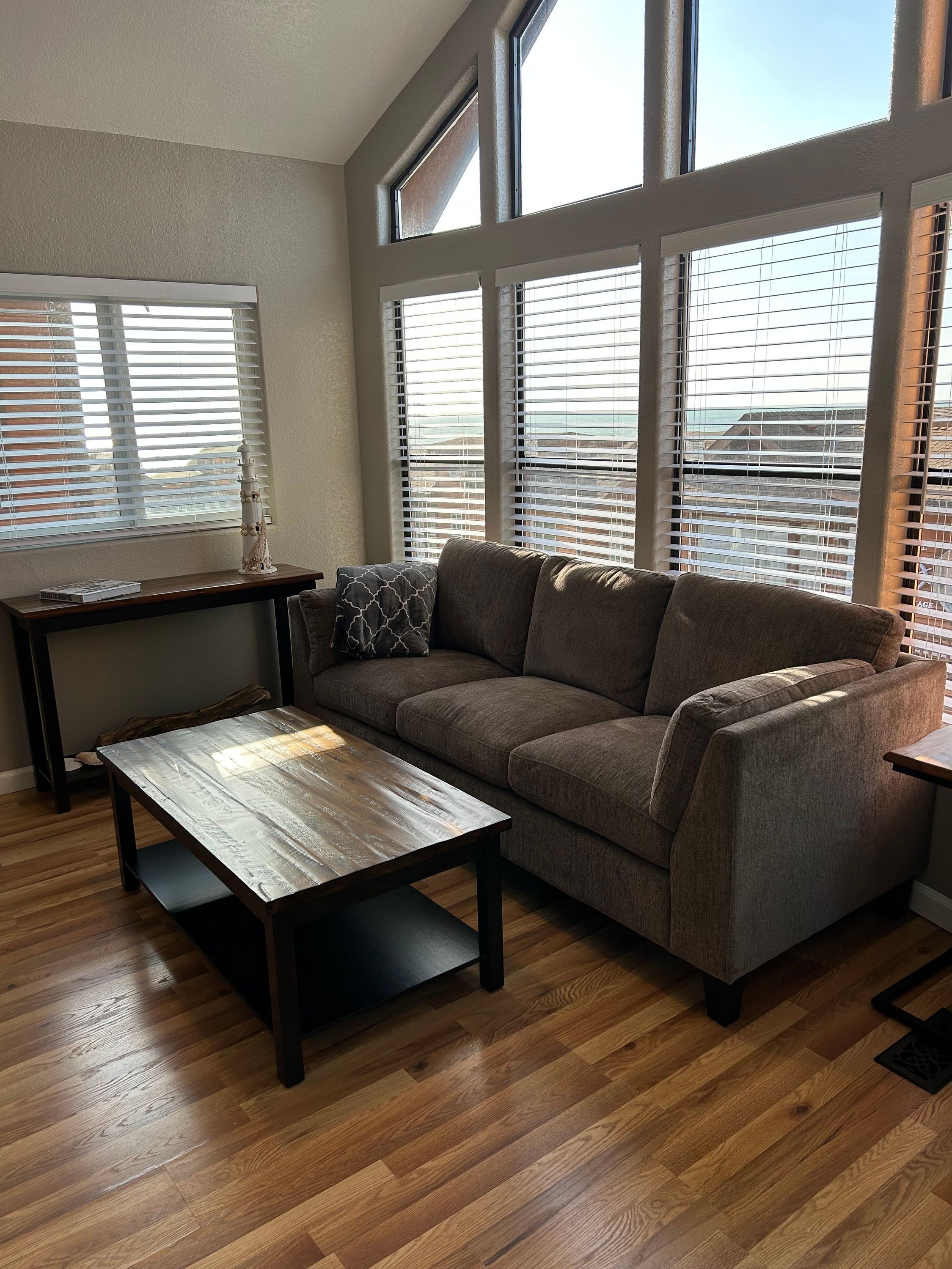 A modern living room with a sectional sofa, a wooden coffee table, and a side table. Large windows with blinds provide natural light and a view of the cityscape.