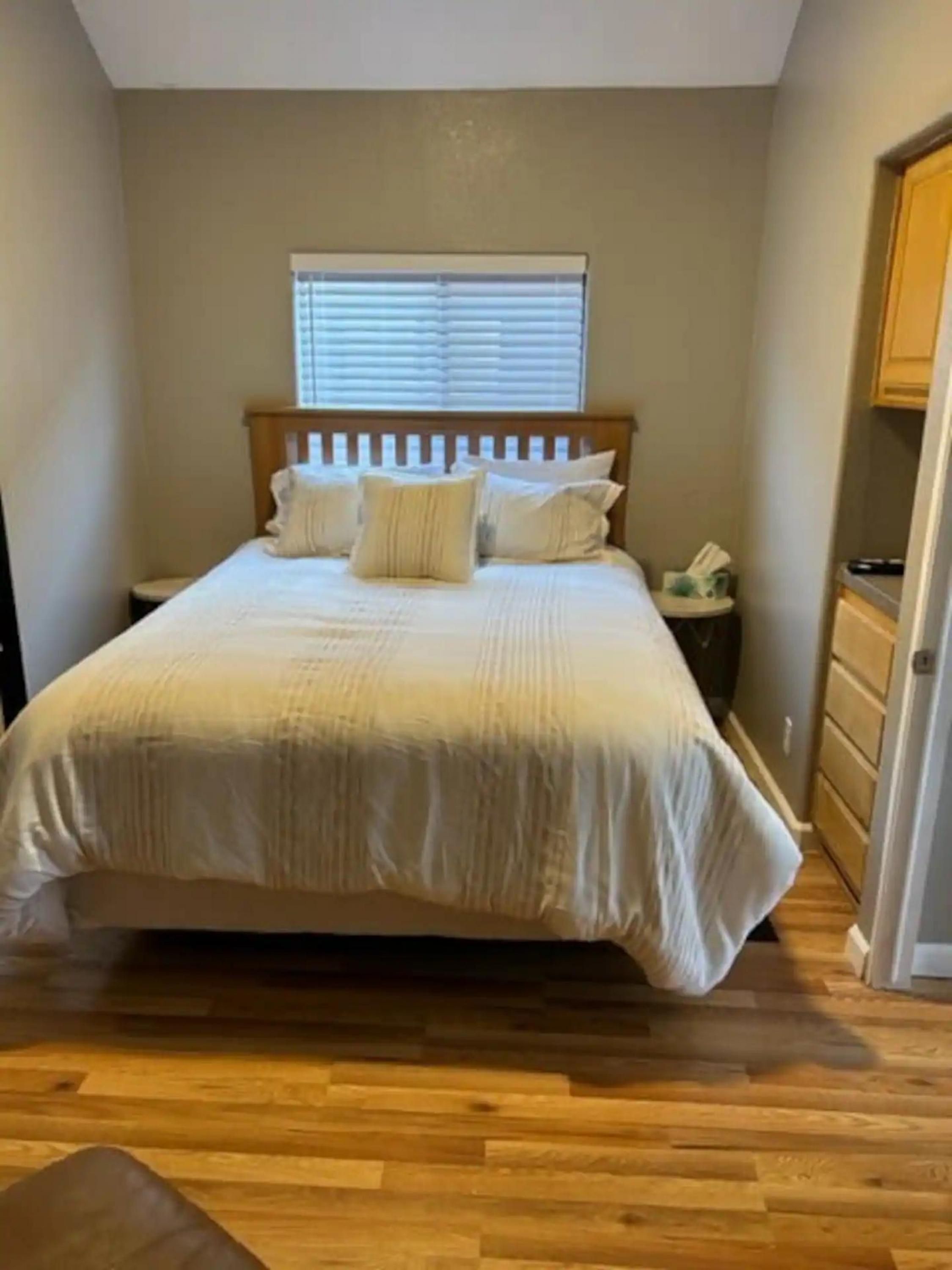 A bedroom with a wooden bed, a nightstand, a window with blinds, and a wooden floor.