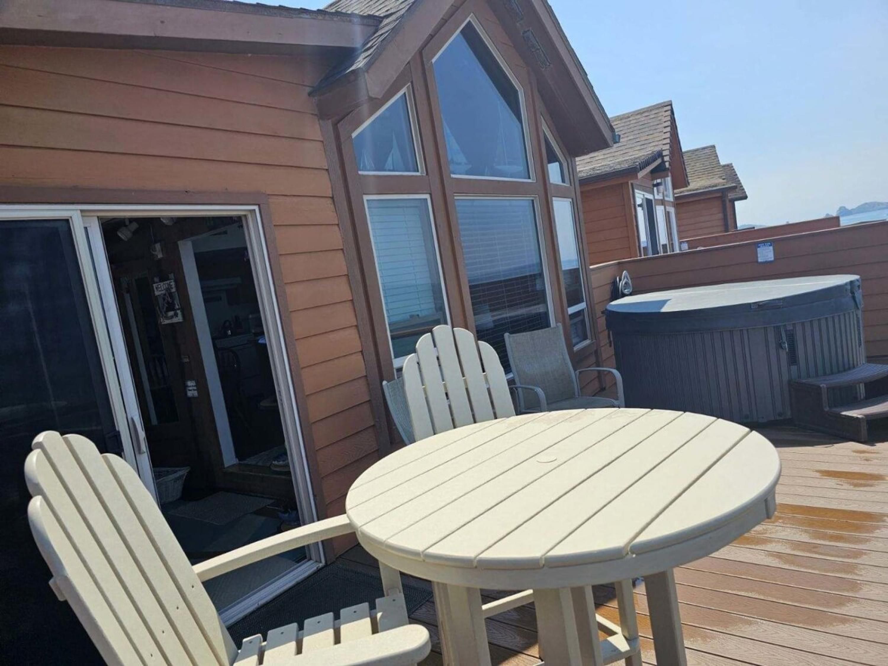 Een terras met een ronde tafel en twee Adirondack-stoelen, een hottub, en een houten huis met grote ramen.