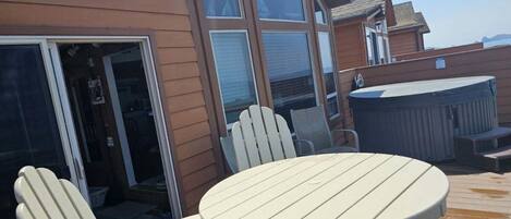 Een terras met een ronde tafel en twee Adirondack-stoelen, een hottub, en een houten huis met grote ramen.