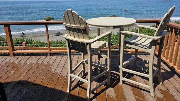 Une terrasse en bois avec deux fauteuils Adirondack et une petite table ronde, donnant sur une plage où l’on peut admirer les vagues et un ciel dégagé.