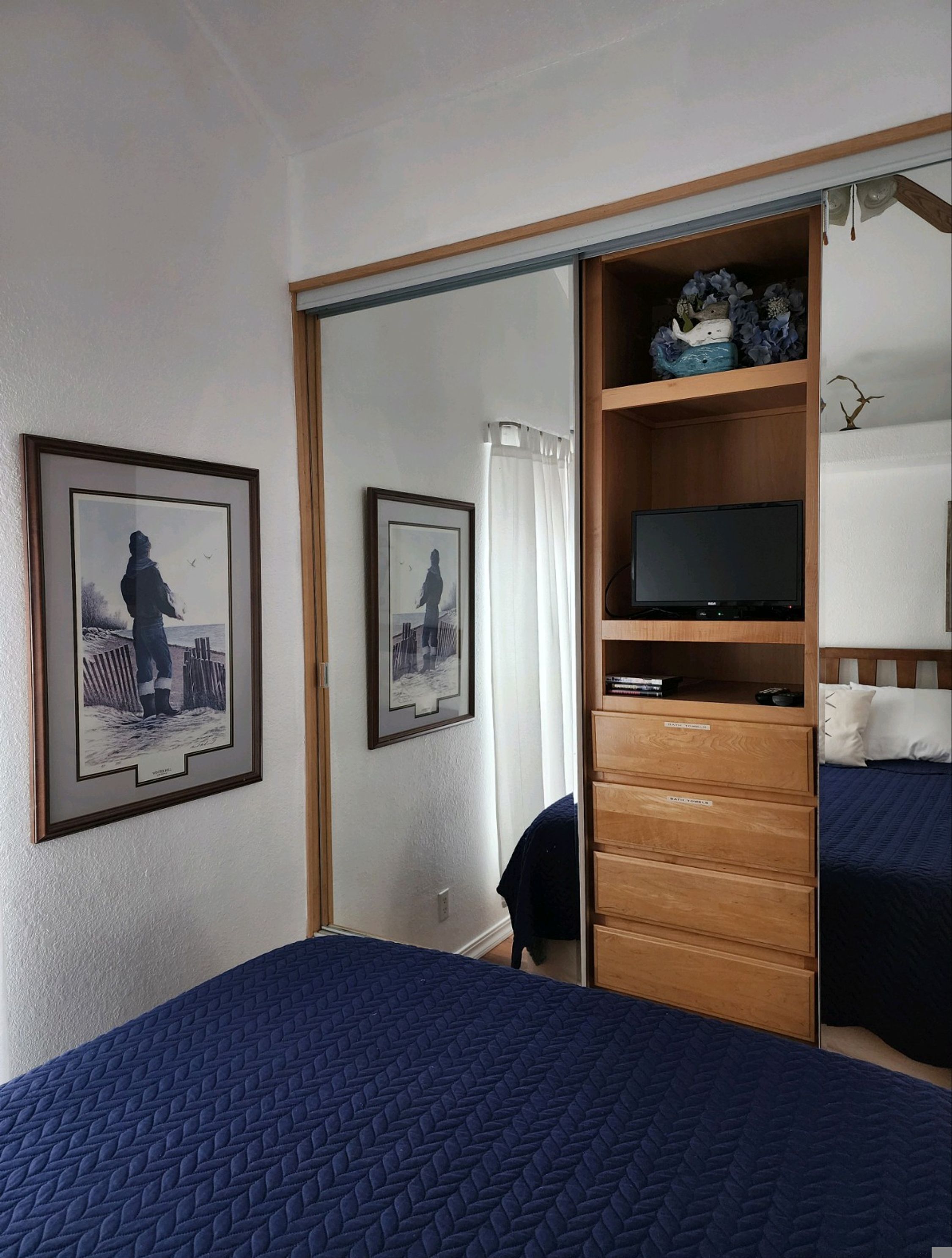 A bedroom with a bed, a TV on a wooden stand, a wardrobe, and two framed pictures on the wall.