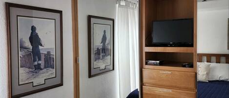 A bedroom with a bed, a TV on a wooden stand, a wardrobe, and two framed pictures on the wall.