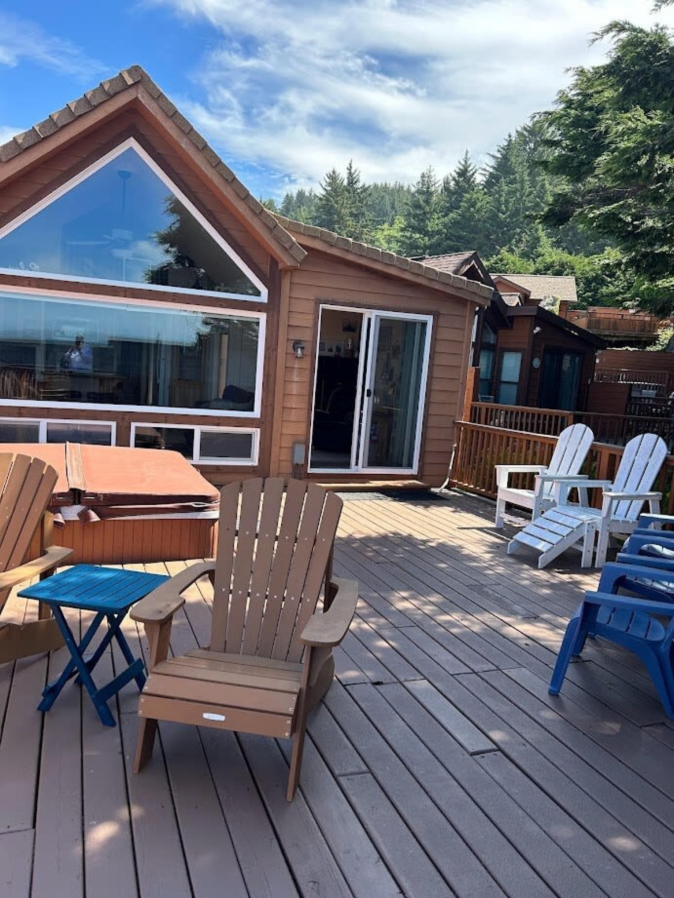 A wooden deck with Adirondack chairs and a table, a glass door entrance, and a view of a forested area.