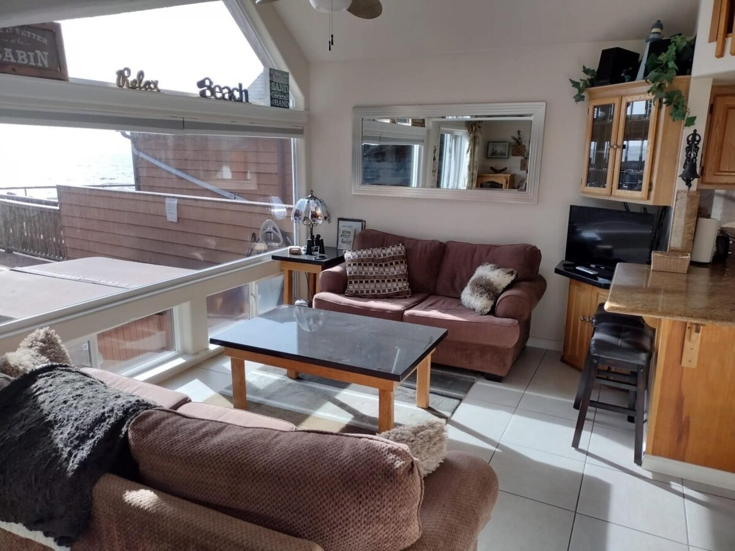 A cozy living room with a sofa, a coffee table, and a TV. There is a balcony with a view of the sea.