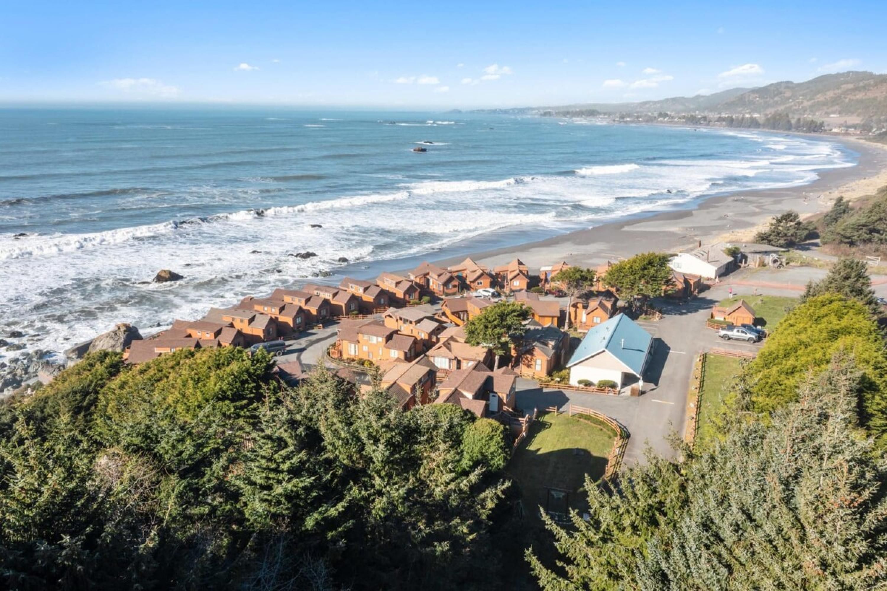 A coastal residential area with houses near the beach, a clear view of the ocean, and a hilly landscape in the background.