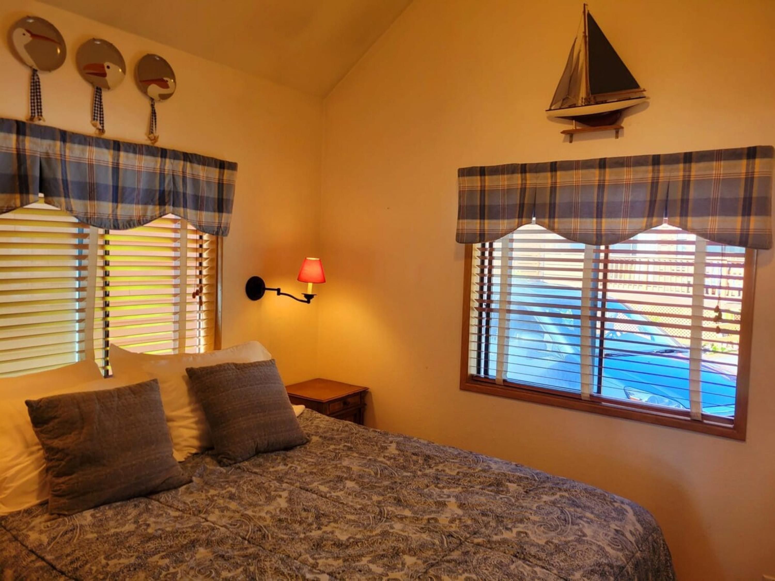 A bedroom with a bed, two pillows, a nightstand, a wall-mounted lamp, and a window with blinds.