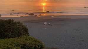 Una playa con un atardecer, costa rocosa y algo de vegetación al frente.