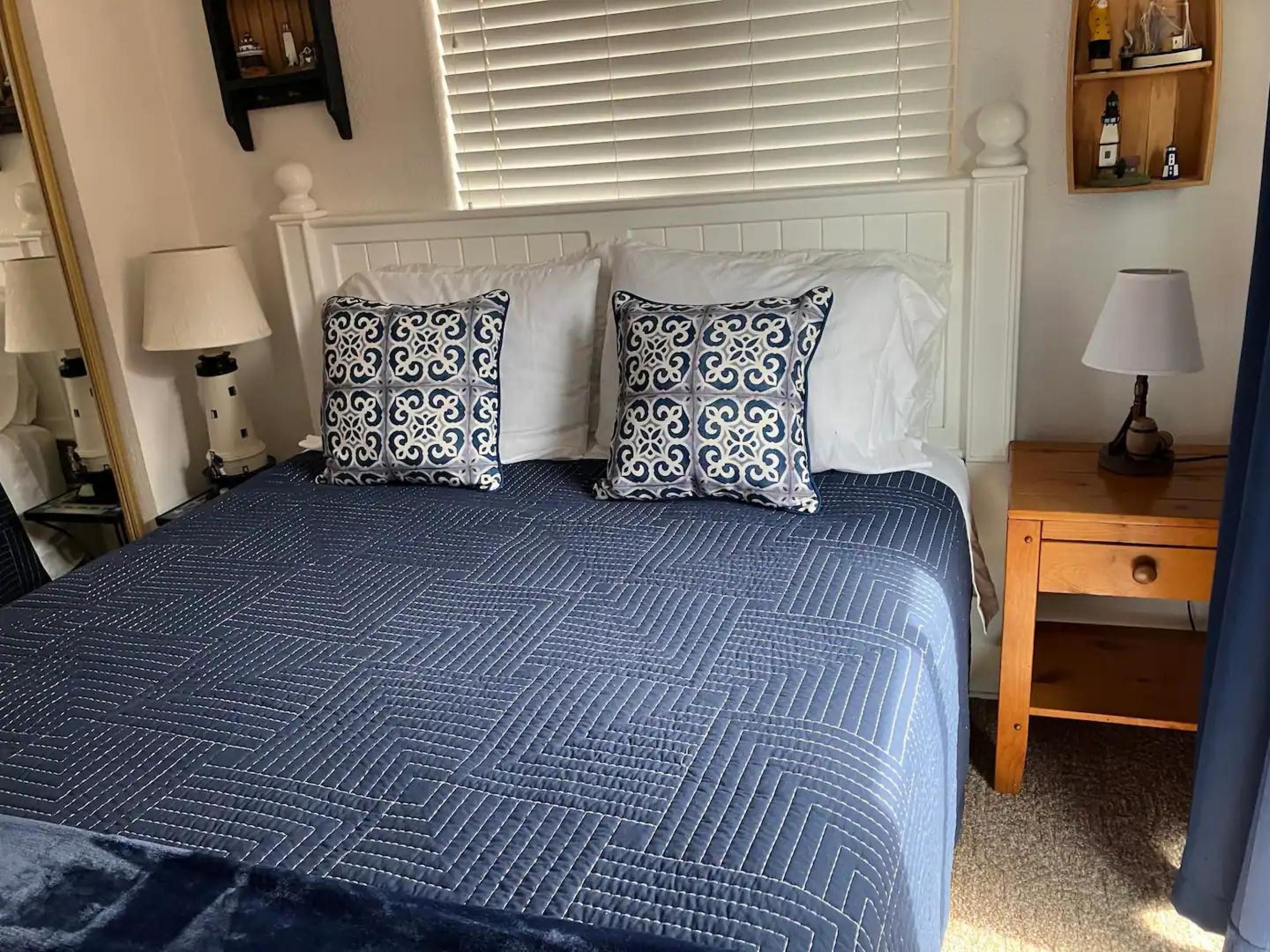 A neatly made bed with a blue quilt and patterned pillows, a bedside table with a lamp, and a shelf with decorative items.