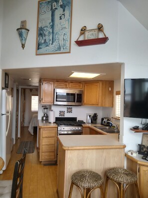 A compact kitchen with wooden cabinets, a stainless steel refrigerator, a microwave, a stove, and a small bar area with stools.