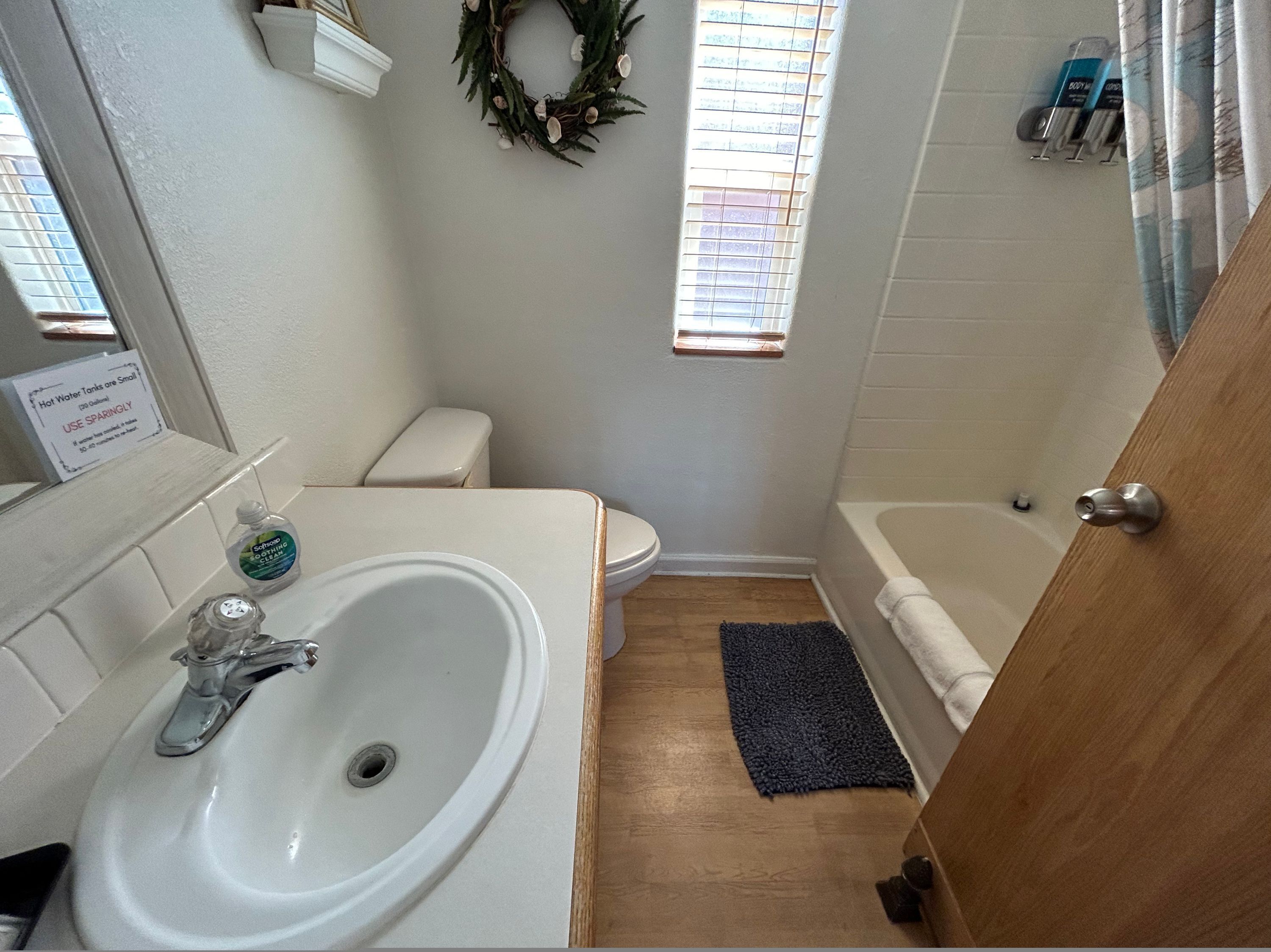 A small bathroom with a white sink, toilet, and bathtub. There is a small window with blinds, a shower curtain, and a sign on the wall.