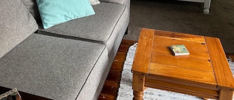 A room with a grey sofa, a wooden coffee table, a bed with a patterned duvet, and a ship's wheel decoration on the wall.