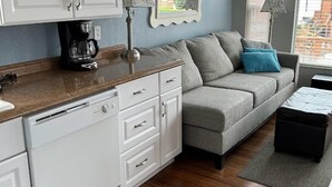 Eine kompakte Küche mit weißen Schränken, einer Kaffeemaschine und einem Essbereich mit grauem Sofa und schwarzem Hocker.