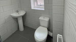 A small, modern bathroom with a white sink, toilet, and a window above the toilet. - Stratford- (London)