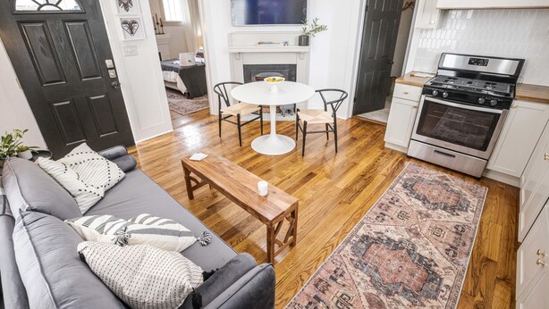 A living room with a gray sofa, a wooden coffee table, and a white round table with chairs. There is a fireplace, a kitchen with a stove, and a large TV mounted above the fireplace. - Evangeline A in Lafayette (Lafayette)