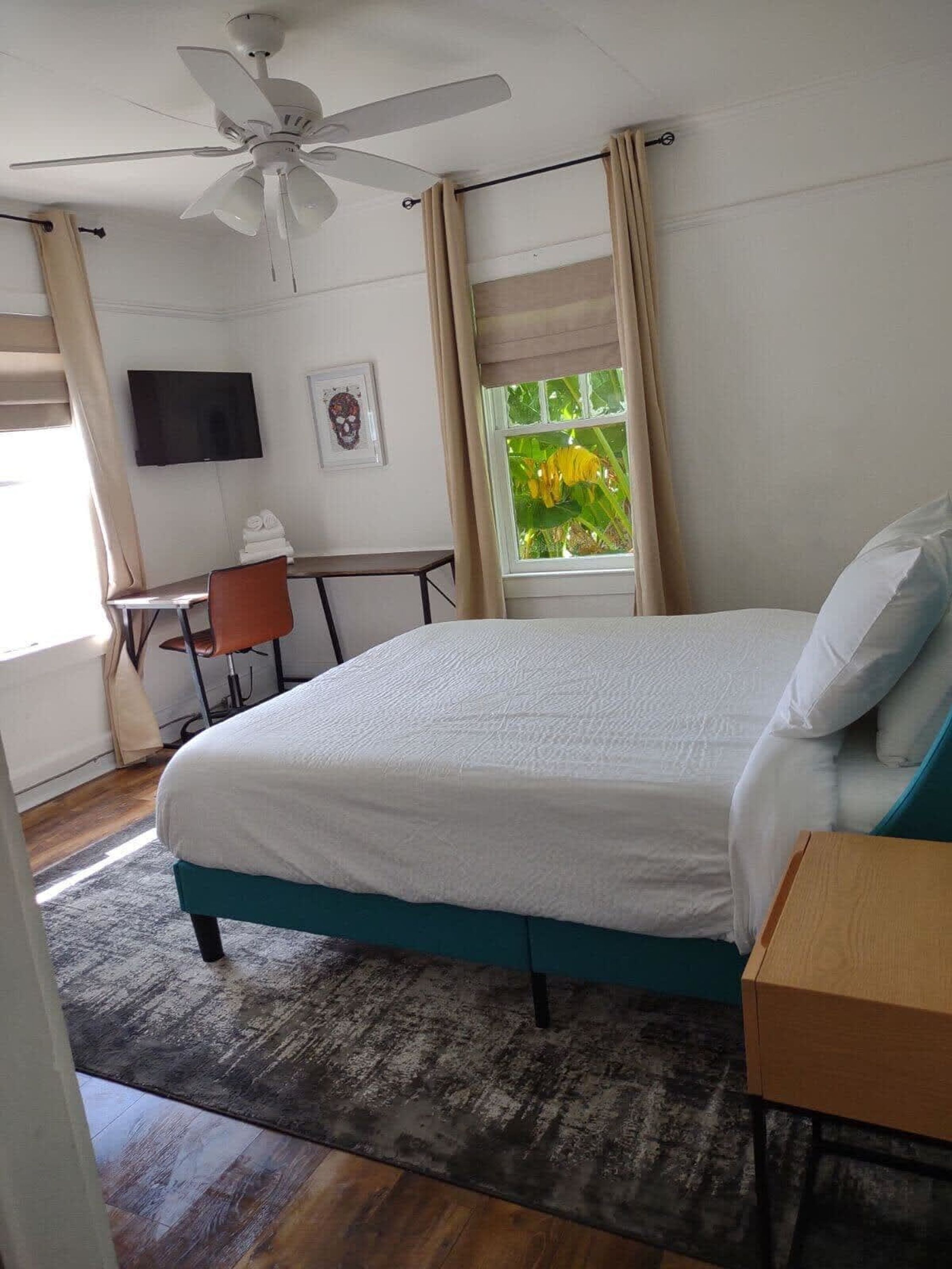A bedroom with a bed, a desk, a chair, a ceiling fan, a TV, and a window with curtains.