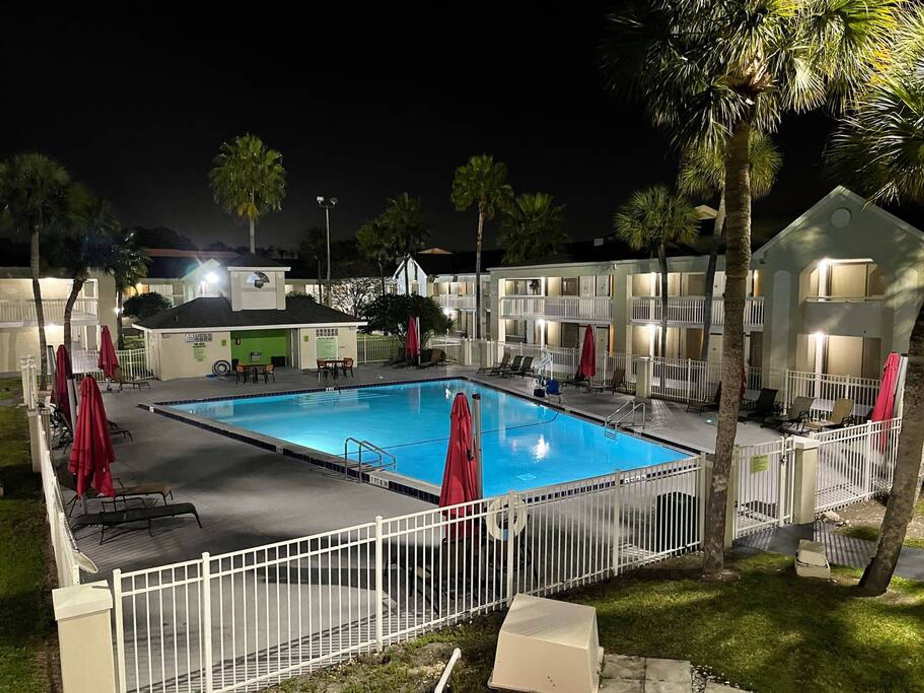 A swimming pool surrounded by a white fence, with palm trees and a building in the background.