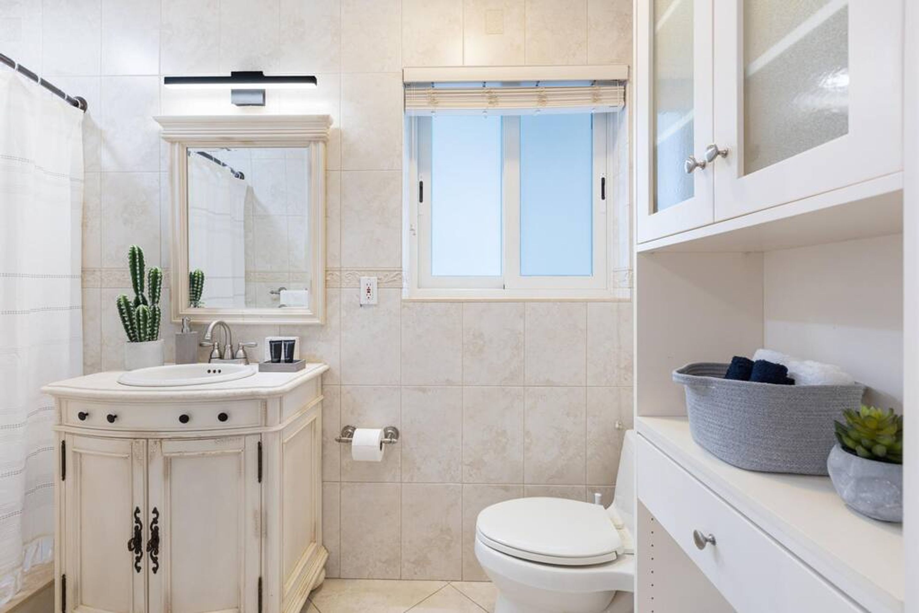 A bathroom with a white sink, mirror, and toilet, a window with blinds, and a cactus decoration.