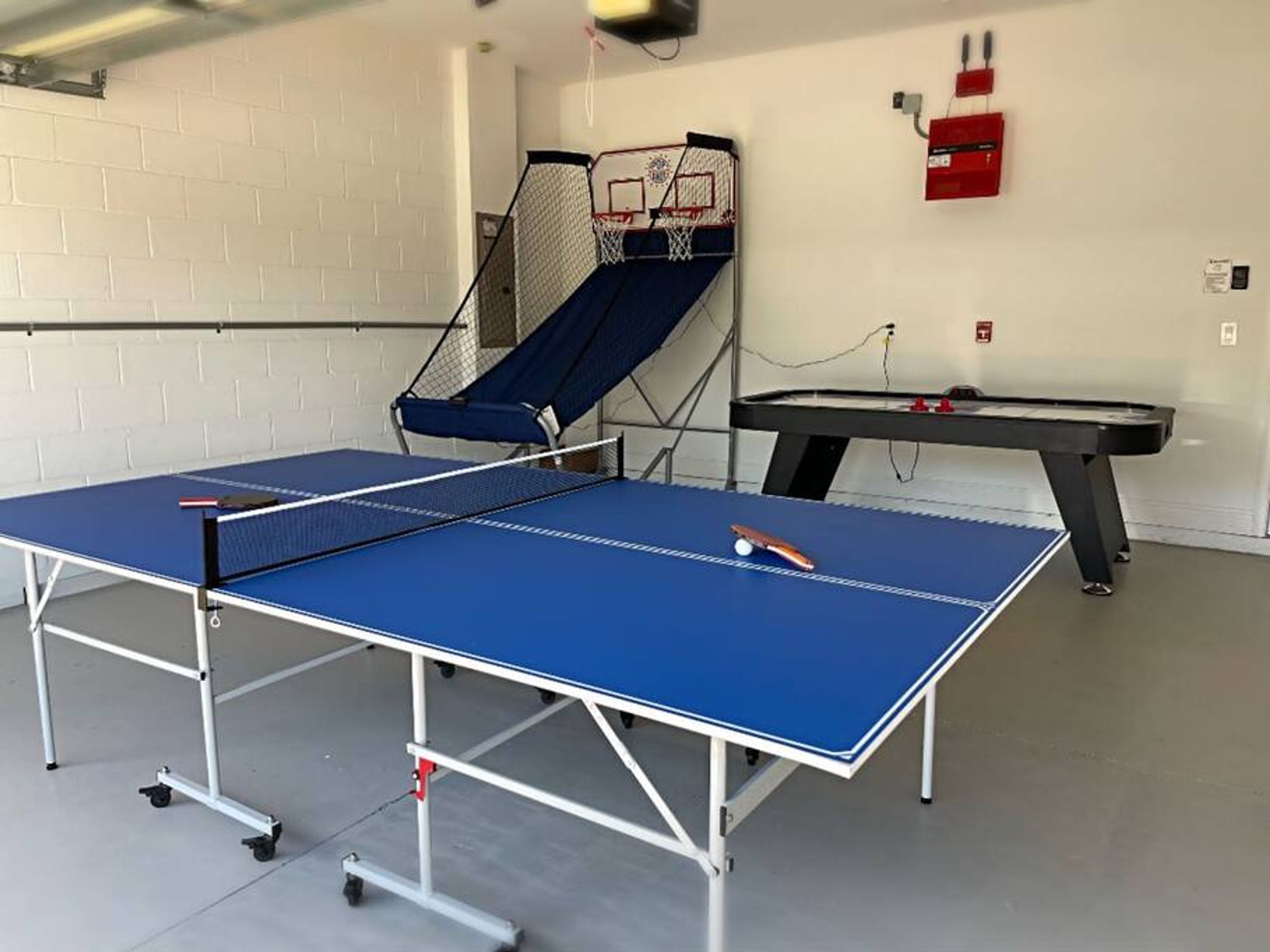 A ping pong table with a net, a basketball hoop, and a pool table in a room with white brick walls.