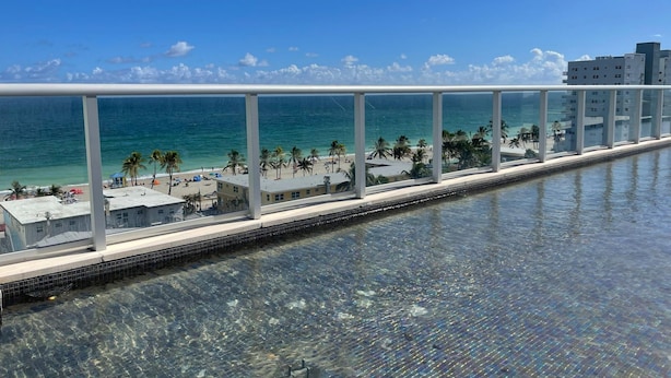 Visão clara de uma piscina com barreira de vidro, uma praia com palmeiras e um céu azul e límpido.