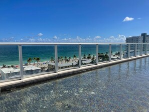 Ein Pool mit Glasbarriere, ein Strand mit Palmen und ein klarer, blauer Himmel sind zu sehen.