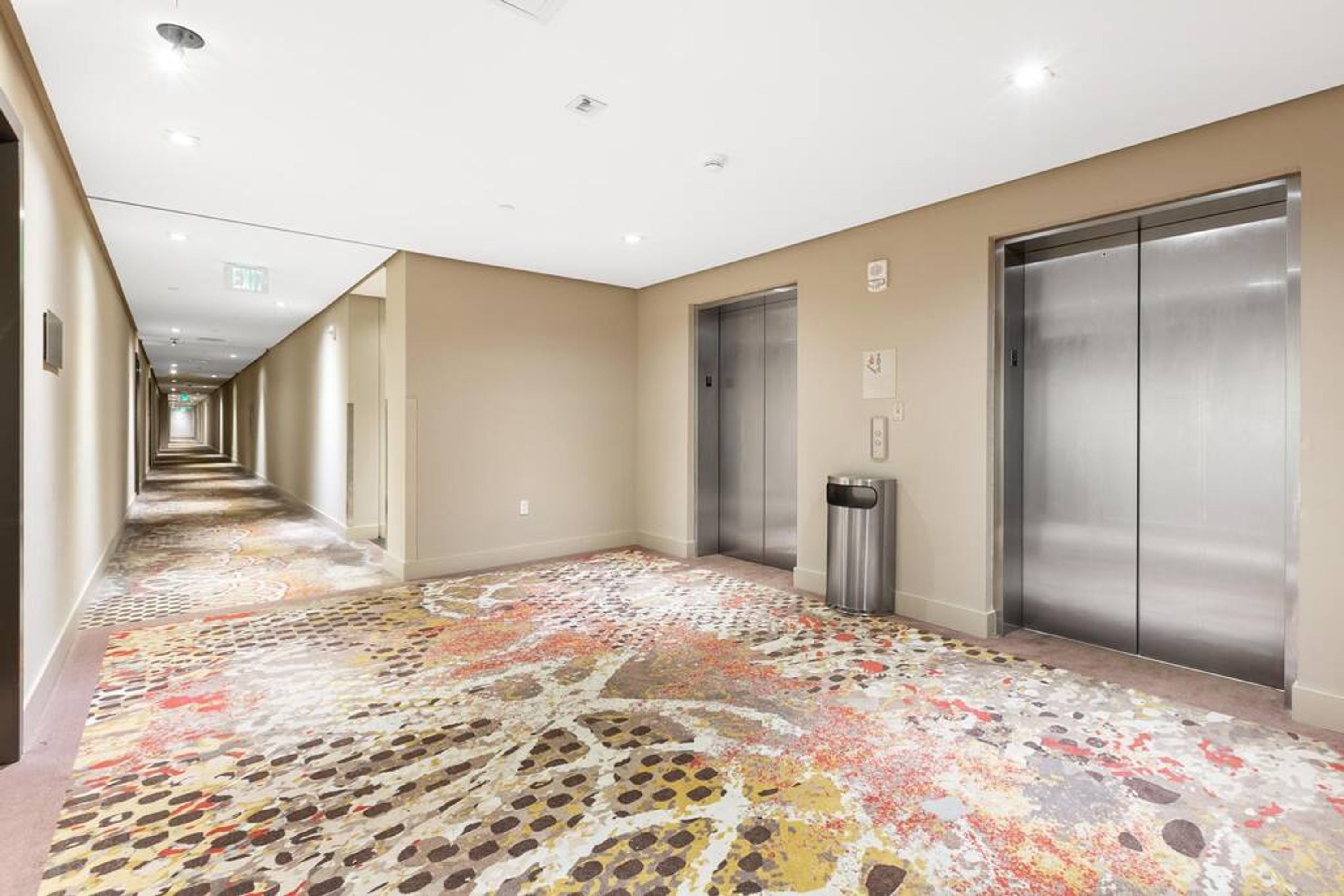 Un couloir d’hôtel avec un tapis à motifs, deux ascenseurs et une poubelle.