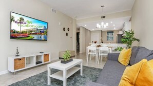 Una sala de estar moderna con un televisor de pantalla plana, un sofá gris con cojines amarillos, una mesa de centro blanca y un comedor con mobiliario blanco.