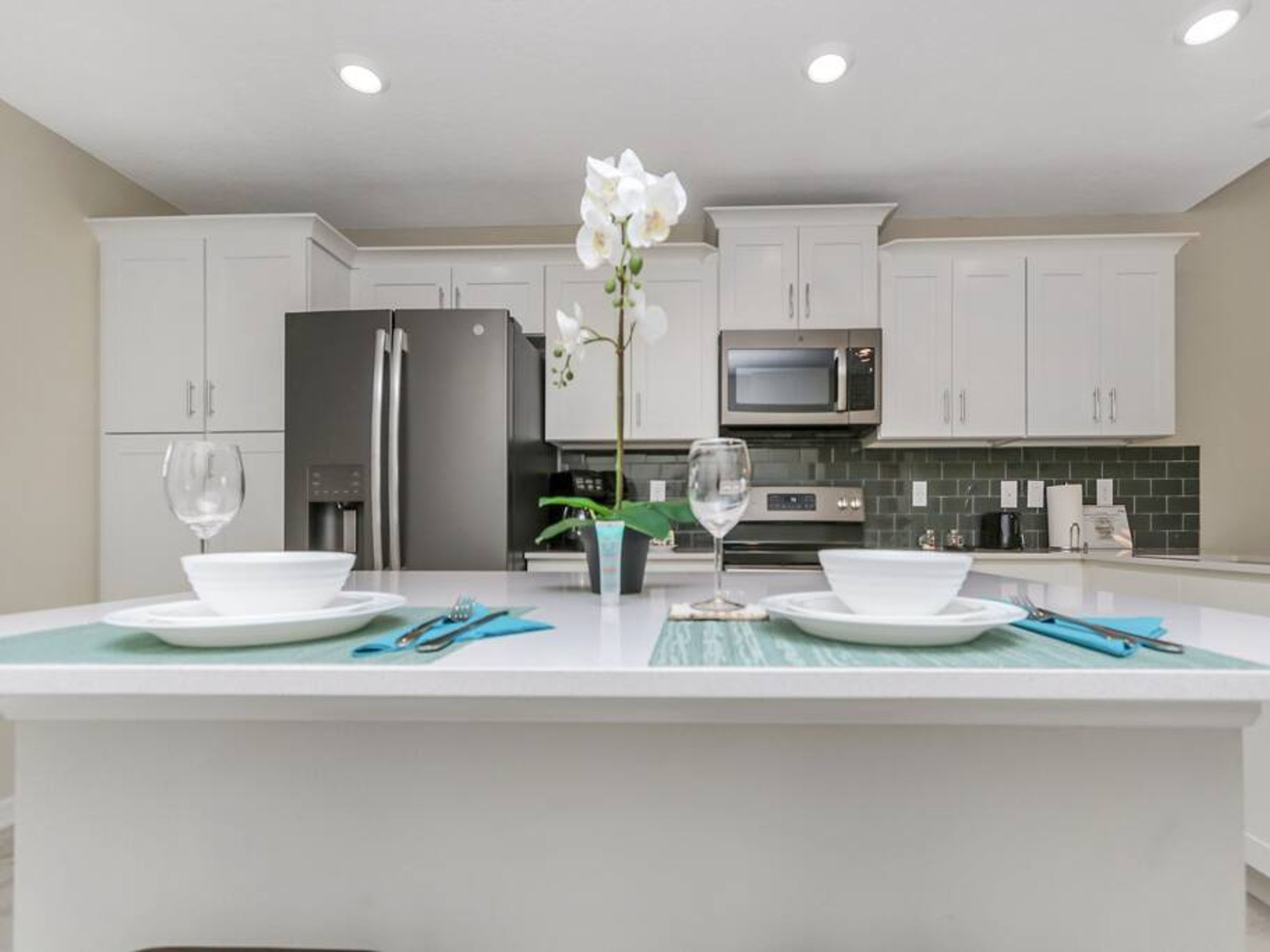 A modern kitchen with white cabinetry, a central island with a vase of white flowers, and a dining setup with plates and glasses.