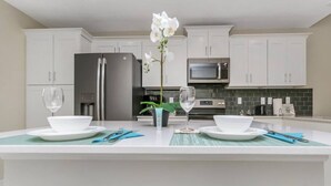 Una cocina moderna con armarios blancos, una isla central con un jarrón de flores blancas y un comedor con platos y vasos.