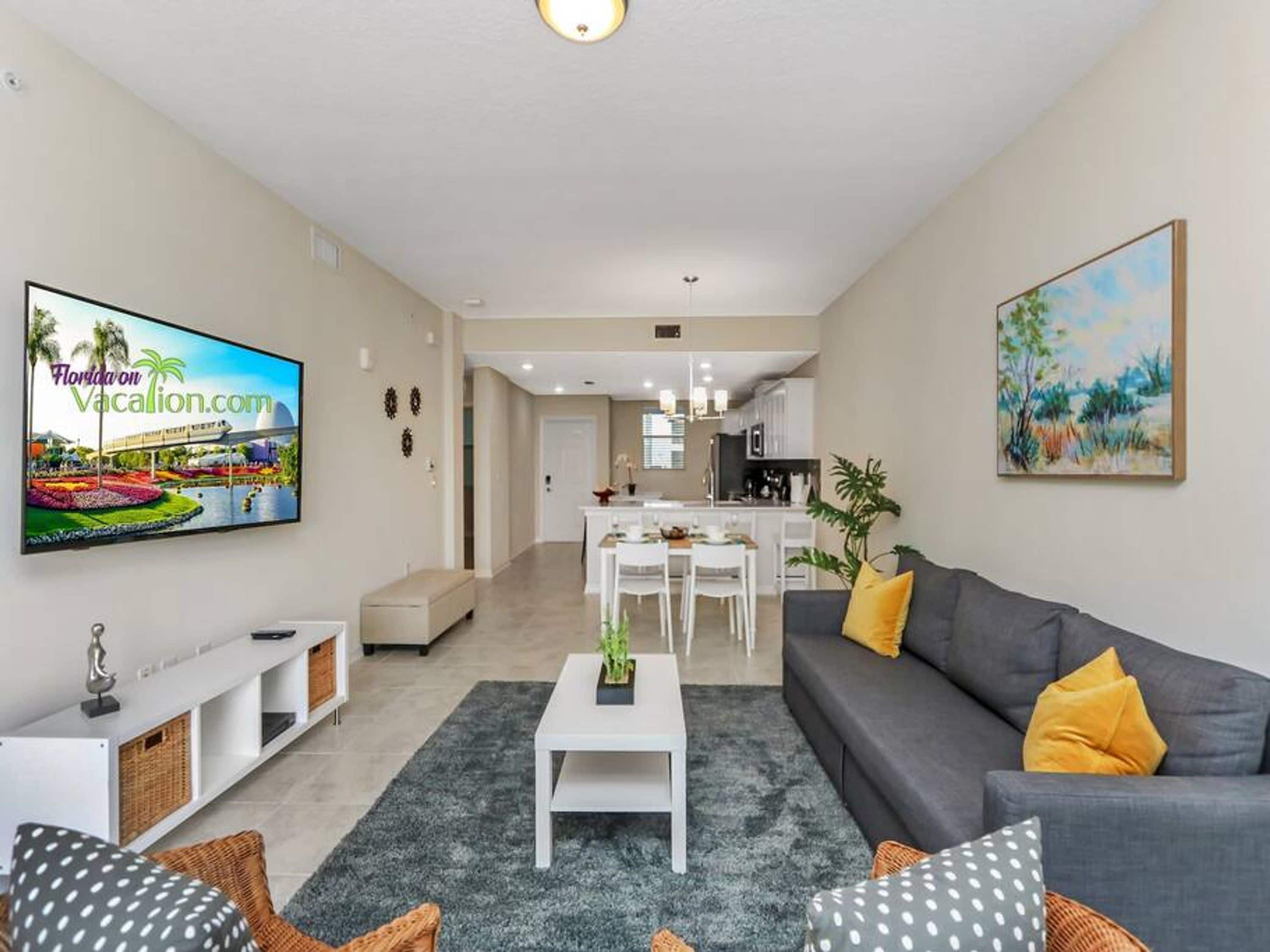 A modern living room with a grey sofa, a white coffee table, and a TV displaying a vacation website. A dining area with a white table and chairs is visible in the background.