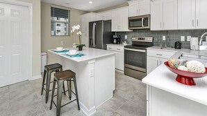 Una cocina moderna con armarios blancos, una isla central con taburetes, electrodomésticos de acero inoxidable y un tazón rojo sobre la encimera.