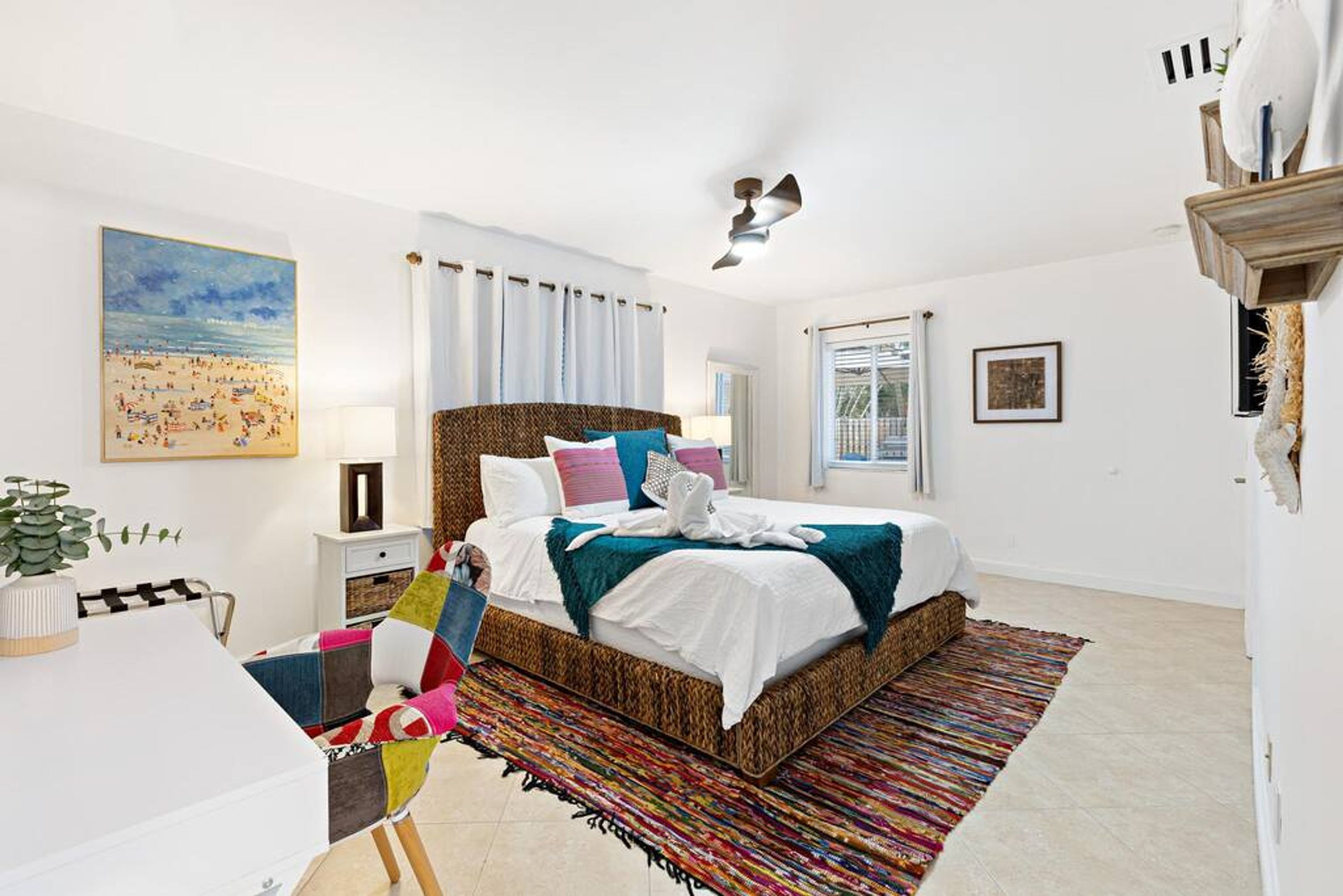 A bedroom with a large bed, a desk, and a chair. There is a window with curtains, a ceiling fan, and a painting on the wall.